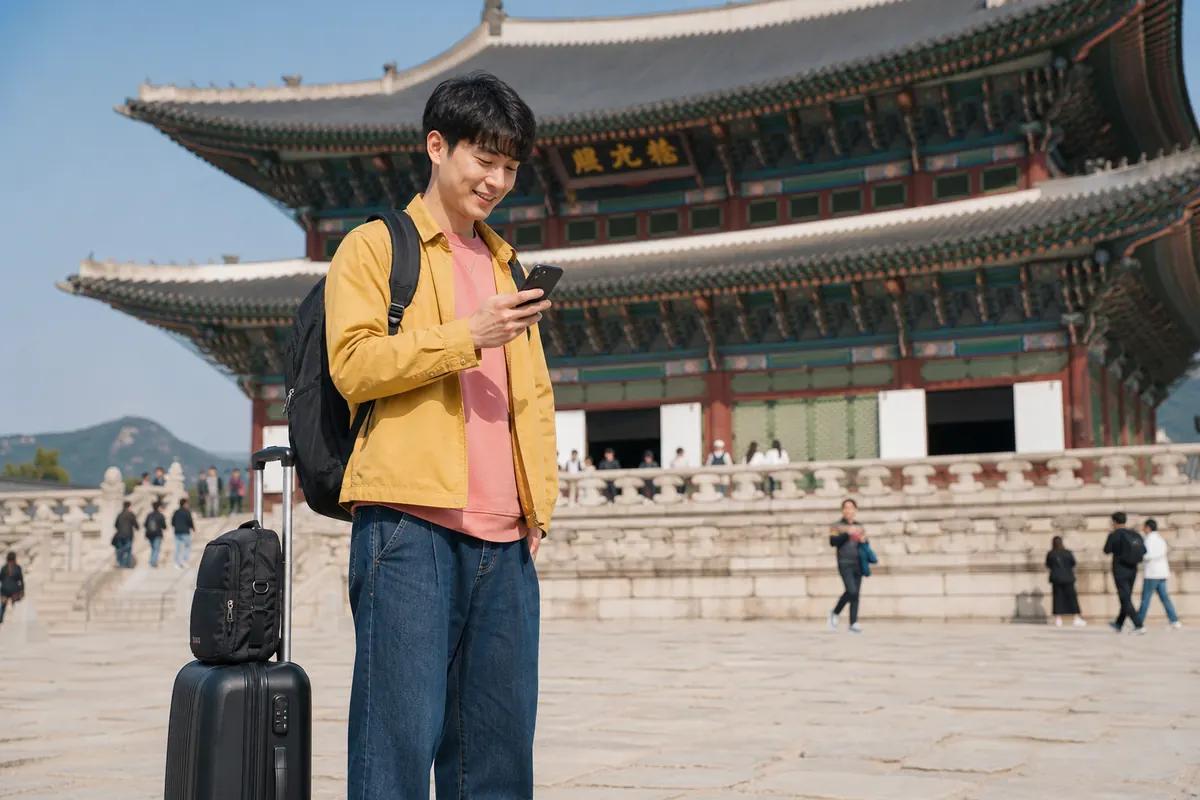 Jeune voyageur souriant sur son smartphone devant un palais traditionnel coréen, valise à ses pieds