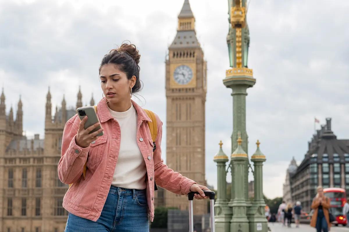 Young woman with a suitcase checking her phone on a bridge, Big Ben rising behind her