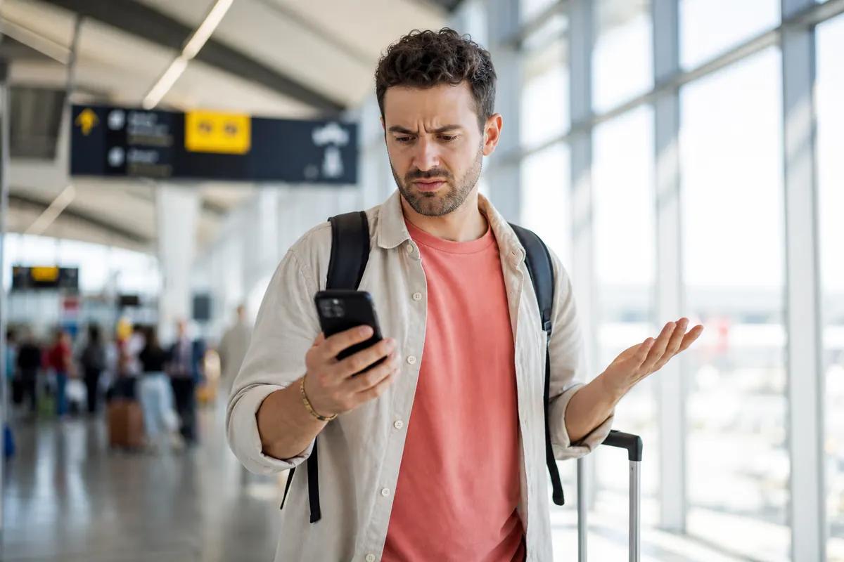 Irritated traveler shrugging at his smartphone screen in a bright airport terminal walkway