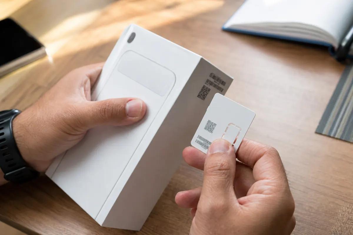 Hands holding a phone box and a SIM card printed with a QR code above a wooden desk