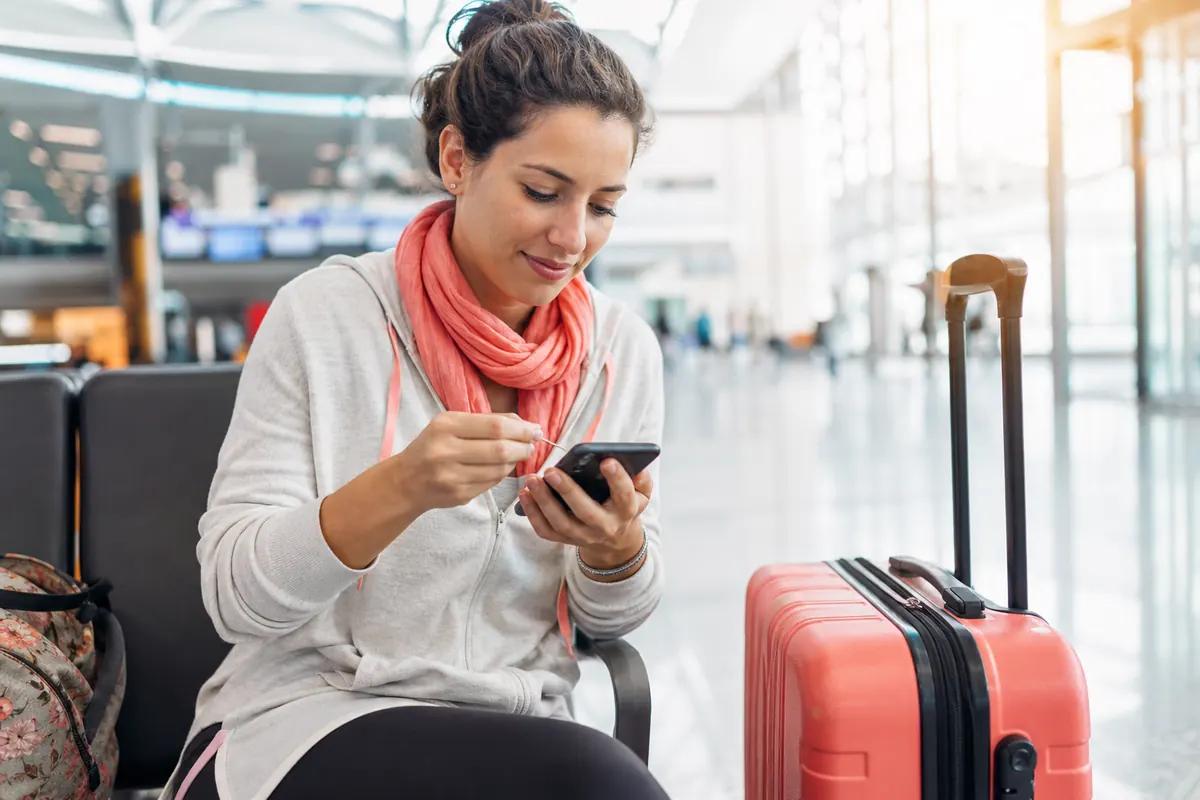 Traveler seated at an airport gate poking a thin pin into her phone's SIM tray, suitcase nearby