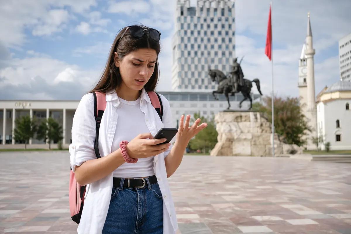 Voyageuse perplexe devant son smartphone sur une place de Tirana, statue équestre en arrière-plan