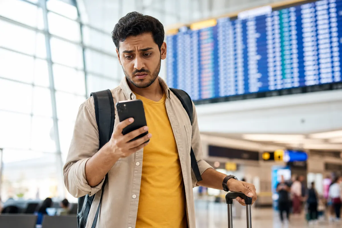 Voyageur inquiet regardant son smartphone devant un panneau d'affichage des vols.